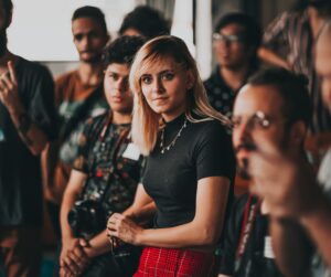 A woman looking ahead wearing a black t-short and red skirt. Other people are in the background but blurred out.